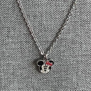 Pode incluir: Um colar prateado com um pingente da Minnie Mouse. O pingente apresenta uma cabe&ccedil;a preta com um la&ccedil;o vermelho e um rosto feito de cristais transparentes. O colar &eacute; exibido em uma superf&iacute;cie cinza texturizada.