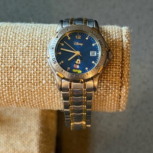 Op de afbeelding: Een zilver- en goudkleurig Disney-horloge met een blauwe wijzerplaat met Mickey Mouse. Het horloge heeft een datumweergave op 3 uur en een roestvrijstalen band. Het horloge wordt getoond op een beige stof.