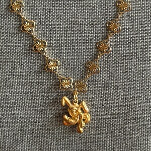 May include: A gold-toned necklace featuring a chain composed of repeating four-petaled flower links with a textured, starburst-like center. A small, three-dimensional gold-toned rabbit pendant hangs from the center of the chain.