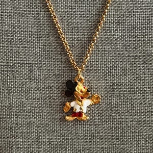 K&ouml;nnte beinhalten: Goldfarbene Halskette mit einem Mickey Mouse-Anh&auml;nger. Der Anh&auml;nger ist ein farbenfrohes Emaille-Design von Mickey Mouse in einer wei&szlig;en Jacke, roten Shorts und schwarzen Ohren. Die Kette ist im filigranen Gliederstil gehalten.