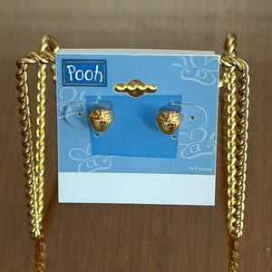 May include: A pair of gold-toned Winnie the Pooh honey pot earrings attached to a light blue display card. The card features the text Pooh and Disney with white line drawings of bees. The earrings are shaped like small pots with the word hunny.