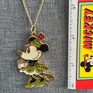 May include: A gold chain necklace with a gold-toned Minnie Mouse pendant. The pendant is green, white and black with red accents. Minnie Mouse is wearing a green dress and hat.