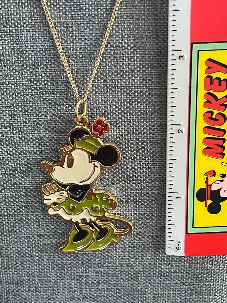 May include: A gold chain necklace with a gold-toned Minnie Mouse pendant. The pendant is green, white and black with red accents. Minnie Mouse is wearing a green dress and hat.