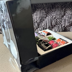 May include: A black box with a Mickey Mouse design on the side, open to reveal a watch and several colorful watch straps. The straps are in various colors, including red, orange, and green. The watch has a silver band and a silver face.