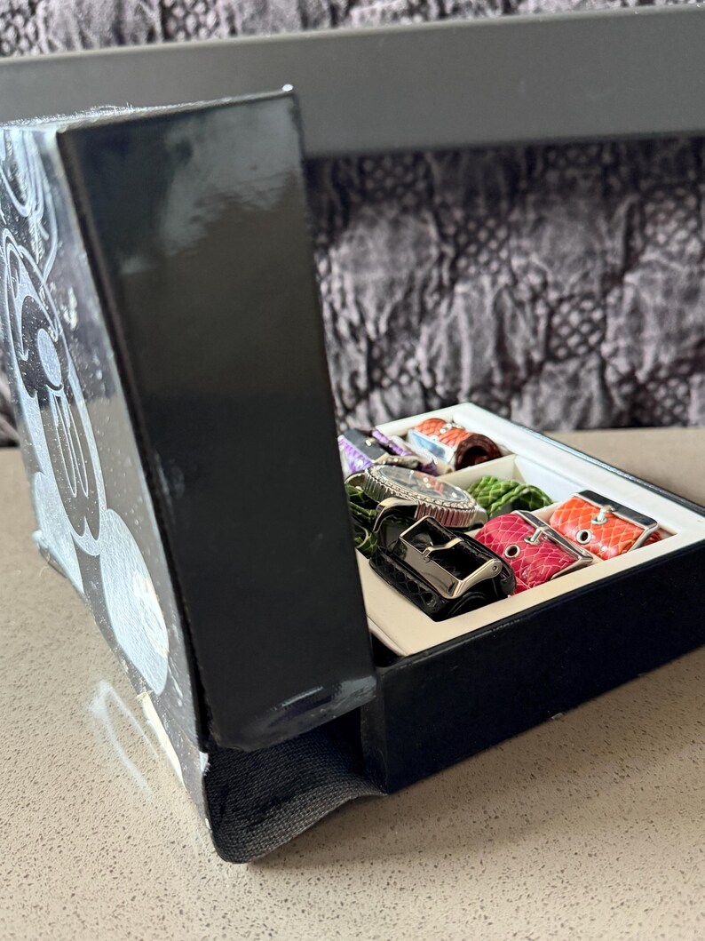 May include: A black box with a Mickey Mouse design on the side, open to reveal a watch and several colorful watch straps. The straps are in various colors, including red, orange, and green. The watch has a silver band and a silver face.