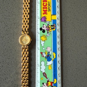 May include: A gold-coloured wristwatch featuring a Mickey Mouse design on the face. The watch has a linked strap and is next to a ruler with Mickey Mouse characters and the text "MICKEY SPORT CLUB".