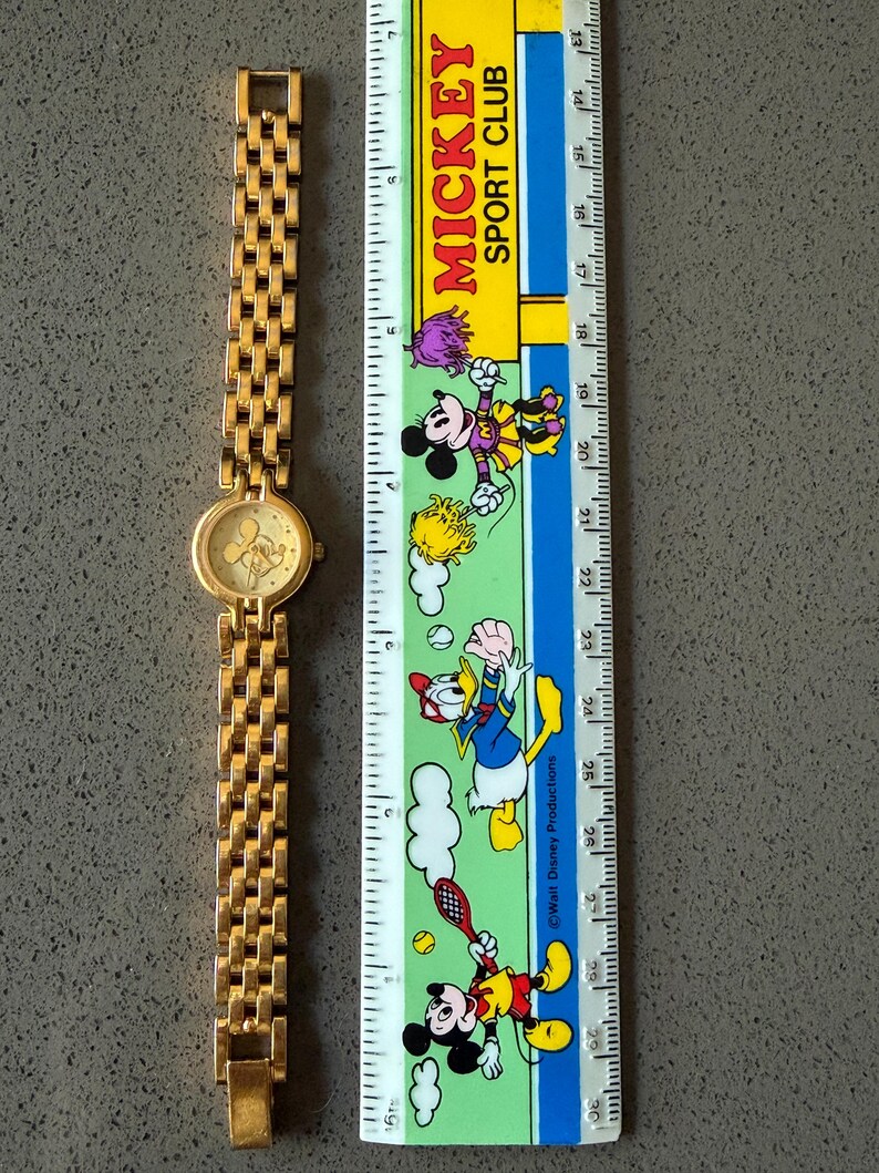 May include: A gold-coloured wristwatch featuring a Mickey Mouse design on the face. The watch has a linked strap and is next to a ruler with Mickey Mouse characters and the text "MICKEY SPORT CLUB".