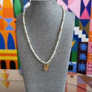 May include: A pearl necklace with gold accents and a gold pendant shaped like a character's head. The necklace is displayed on a gray mannequin against a colorful geometric background.