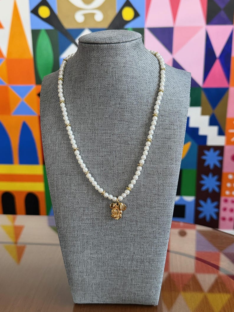 May include: A pearl necklace with gold accents and a gold pendant shaped like a character's head. The necklace is displayed on a gray mannequin against a colorful geometric background.