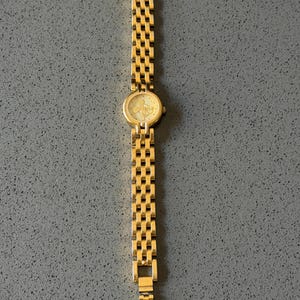 May include: A gold-coloured wristwatch with a rectangular link strap. The watch face is round with a light-coloured background and a Mickey Mouse design. The watch is displayed on a grey surface.