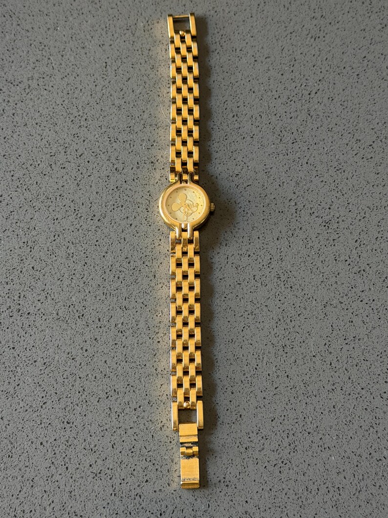 May include: A gold-coloured wristwatch with a rectangular link strap. The watch face is round with a light-coloured background and a Mickey Mouse design. The watch is displayed on a grey surface.