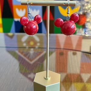 Puede incluir: Un par de pendientes rojos con forma de Mickey Mouse en un soporte dorado. Los pendientes est&aacute;n hechos de un material brillante y liso.