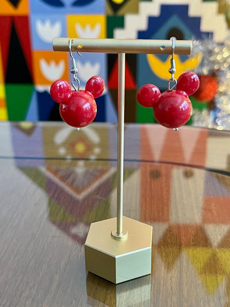 Puede incluir: Un par de pendientes rojos con forma de Mickey Mouse en un soporte dorado. Los pendientes est&aacute;n hechos de un material brillante y liso.