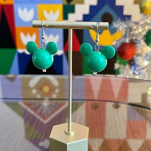 Puede incluir: Un par de pendientes verdes con forma de cabeza de Mickey Mouse en un soporte dorado. Los pendientes son de pl&aacute;stico y tienen ganchos plateados.