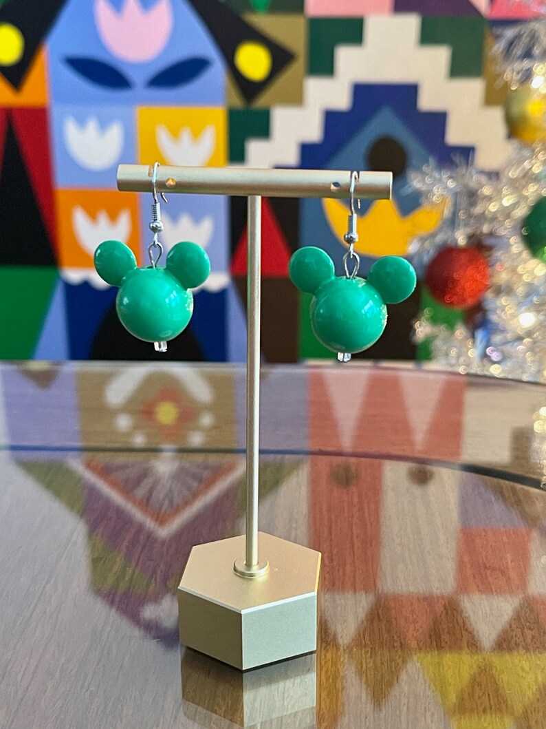 Puede incluir: Un par de pendientes verdes con forma de cabeza de Mickey Mouse en un soporte dorado. Los pendientes son de pl&aacute;stico y tienen ganchos plateados.
