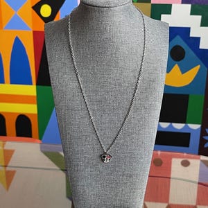 Pode incluir: Colar prateado com um pingente em forma de cabe&ccedil;a de rato. O pingente tem detalhes vermelhos e brancos. O colar &eacute; exibido em um manequim cinza sobre um fundo colorido.