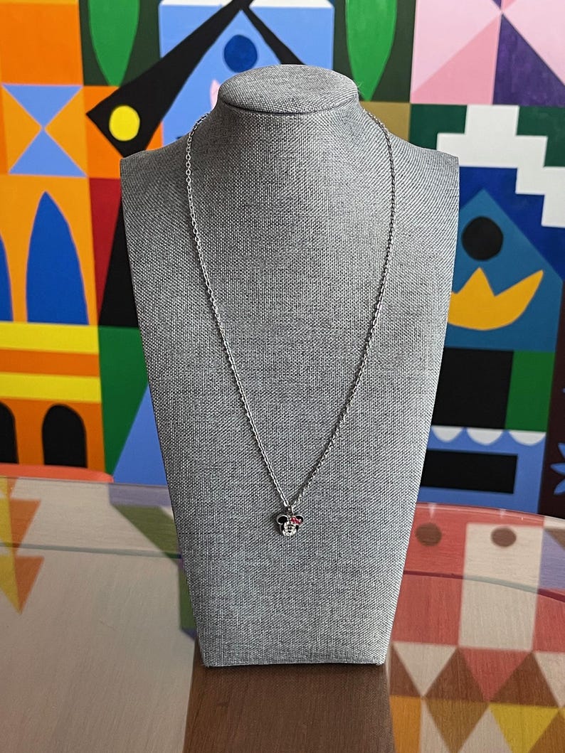 Pode incluir: Colar prateado com um pingente em forma de cabe&ccedil;a de rato. O pingente tem detalhes vermelhos e brancos. O colar &eacute; exibido em um manequim cinza sobre um fundo colorido.