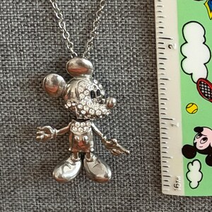 May include: Silver Mickey Mouse pendant necklace. The pendant depicts Mickey Mouse with rhinestone detailing on the face. The necklace is shown on a grey surface, with a ruler and a colourful sticker in the background.