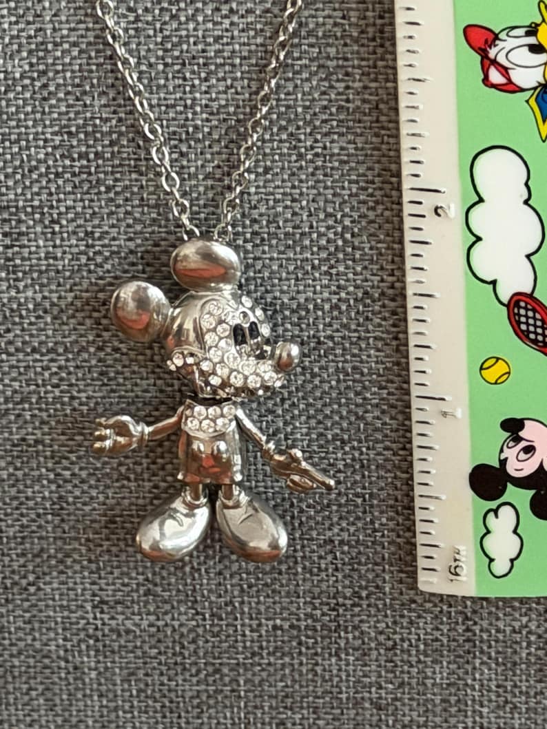 May include: Silver Mickey Mouse pendant necklace. The pendant depicts Mickey Mouse with rhinestone detailing on the face. The necklace is shown on a grey surface, with a ruler and a colourful sticker in the background.
