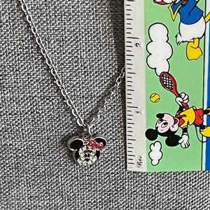 Pode incluir: Um colar prateado com um pingente da Minnie Mouse. O pingente apresenta a silhueta preta da cabe&ccedil;a da Minnie, adornada com um la&ccedil;o vermelho e cristais brilhantes. A corrente &eacute; delicada e o pingente tem cerca de 2 cm de largura.