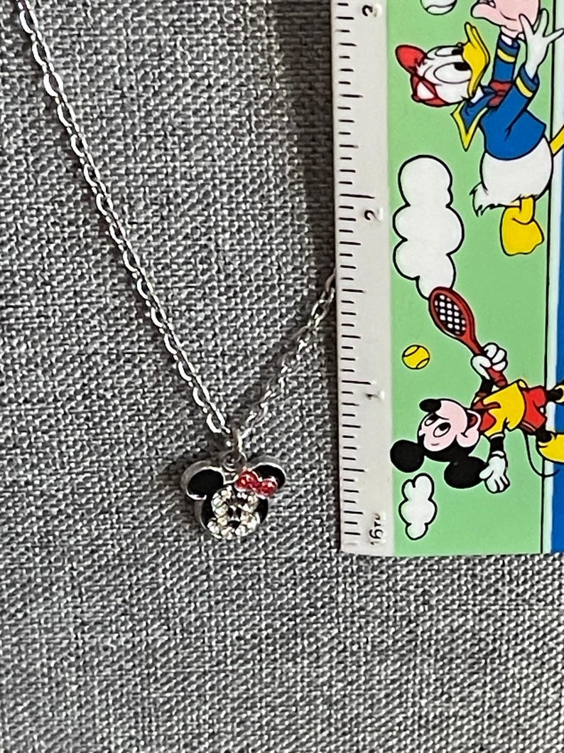 Pode incluir: Um colar prateado com um pingente da Minnie Mouse. O pingente apresenta a silhueta preta da cabe&ccedil;a da Minnie, adornada com um la&ccedil;o vermelho e cristais brilhantes. A corrente &eacute; delicada e o pingente tem cerca de 2 cm de largura.