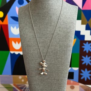 May include: A silver Mickey Mouse pendant necklace on a grey mannequin. The pendant shows a detailed Mickey Mouse figure. The necklace is set against a colourful abstract background.