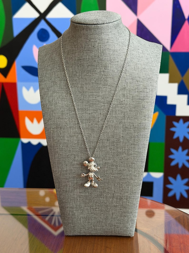 May include: A silver Mickey Mouse pendant necklace on a grey mannequin. The pendant shows a detailed Mickey Mouse figure. The necklace is set against a colourful abstract background.