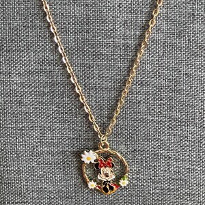 May include: Gold-colored chain necklace with a pendant featuring a cartoon character in a red bow and dress, surrounded by a wreath of white daisies. The pendant is circular and has black, red, and yellow accents.