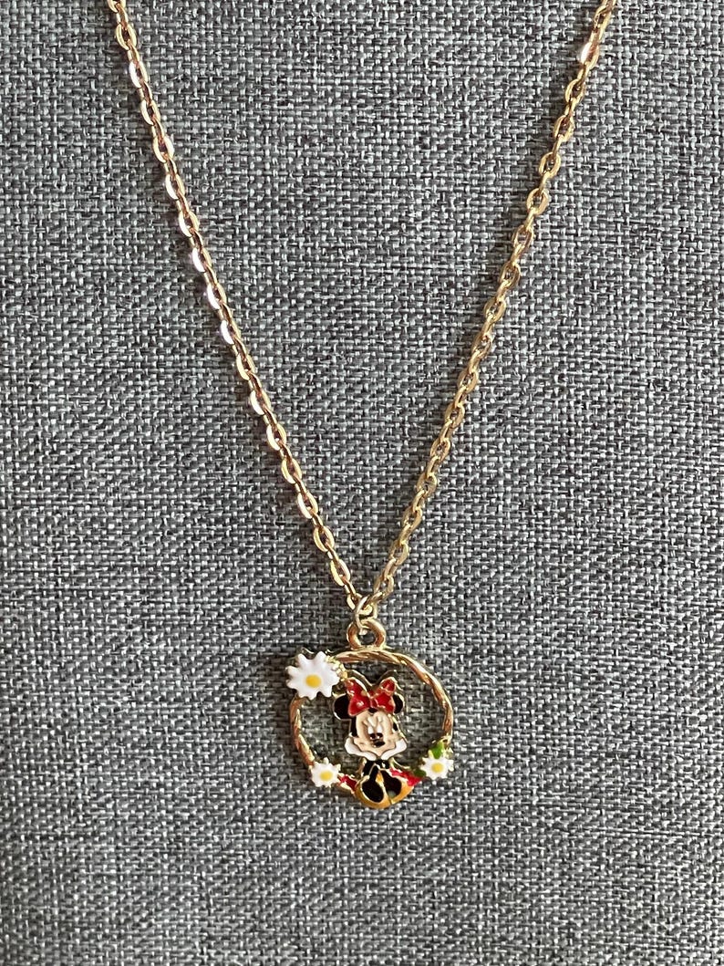 May include: Gold-colored chain necklace with a pendant featuring a cartoon character in a red bow and dress, surrounded by a wreath of white daisies. The pendant is circular and has black, red, and yellow accents.