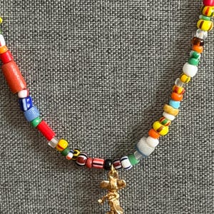 May include: A colorful beaded necklace with a gold-colored charm. The necklace features a variety of small, multi-colored beads, including red, blue, yellow, and green. The charm is a small, detailed figure.