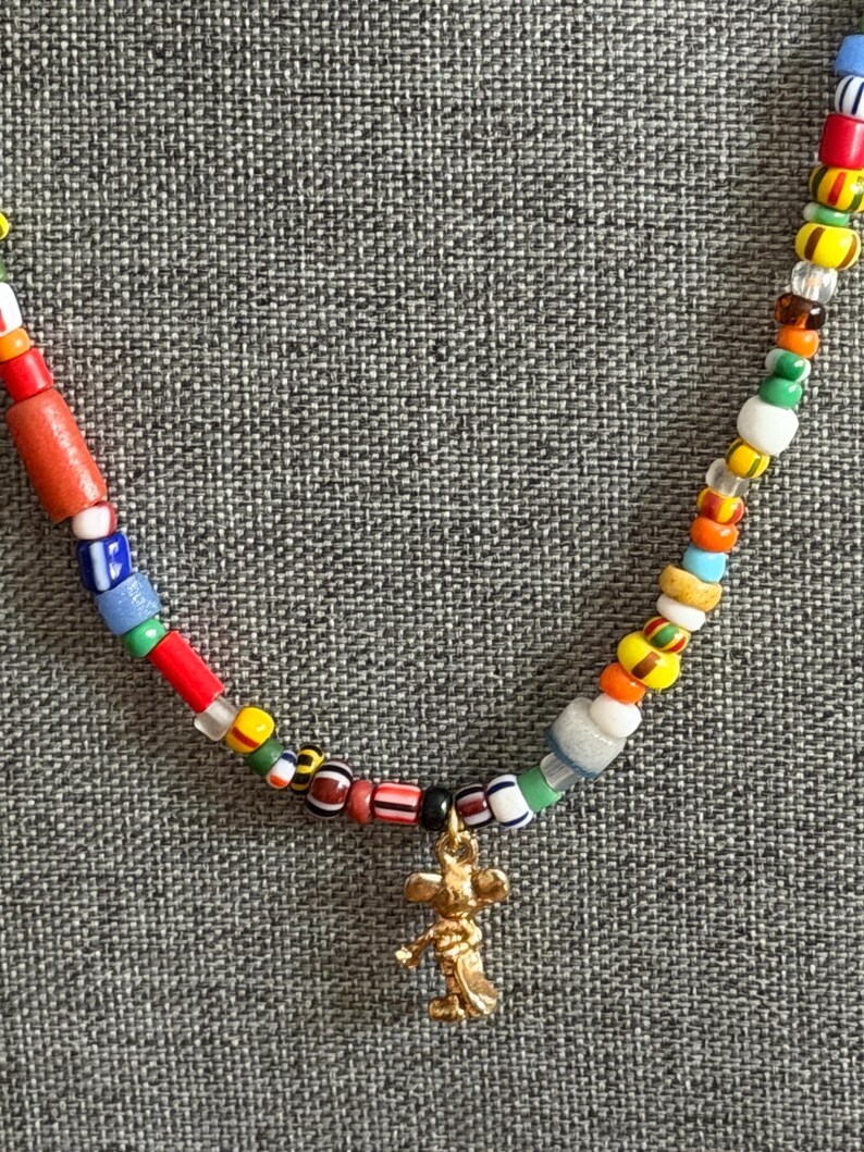 May include: A colorful beaded necklace with a gold-colored charm. The necklace features a variety of small, multi-colored beads, including red, blue, yellow, and green. The charm is a small, detailed figure.