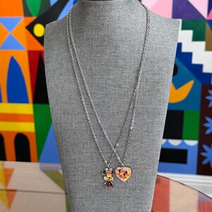 Peut inclure: Deux colliers argent&eacute;s pr&eacute;sent&eacute;s sur un mannequin gris. L'un comporte un pendentif Minnie Mouse, l'autre un pendentif en forme de c&oelig;ur avec un motif Mickey et Minnie Mouse. Les pendentifs ont des accents color&eacute;s.