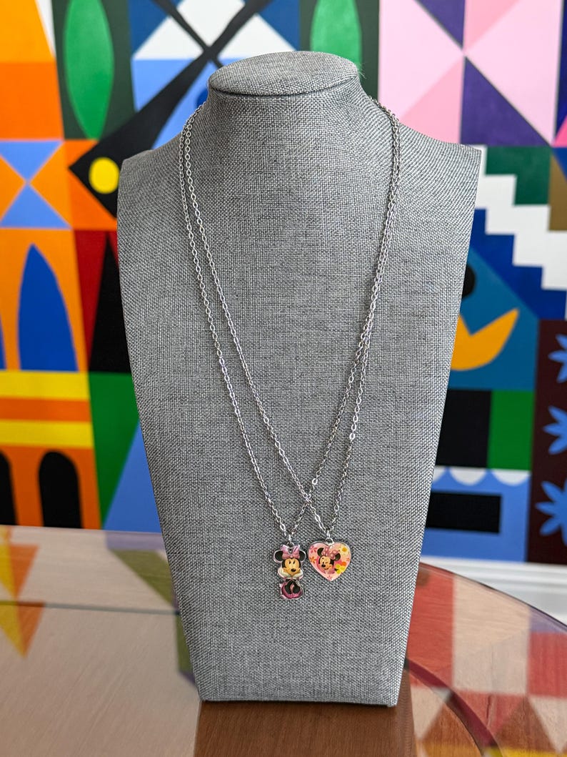 Peut inclure: Deux colliers argent&eacute;s pr&eacute;sent&eacute;s sur un mannequin gris. L'un comporte un pendentif Minnie Mouse, l'autre un pendentif en forme de c&oelig;ur avec un motif Mickey et Minnie Mouse. Les pendentifs ont des accents color&eacute;s.