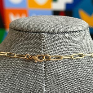 May include: A gold-colored chain necklace with a lobster clasp closure. The necklace is displayed on a gray mannequin head. The background features a colorful abstract design.