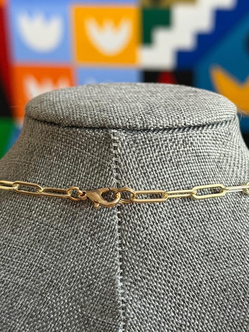 May include: A gold-colored chain necklace with a lobster clasp closure. The necklace is displayed on a gray mannequin head. The background features a colorful abstract design.