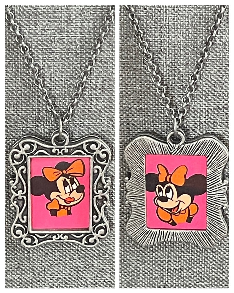 May include: Two silver necklaces featuring a framed image of Minnie Mouse. The image is set against a pink background with orange accents. The necklaces are displayed on a grey fabric background.
