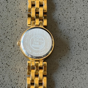 May include: A gold-coloured Disney Time Works watch with a round face and a linked strap. The watch face has a silver backing with the Disney Time Works logo and the text "Thailand Movt". The strap is made of rectangular links.