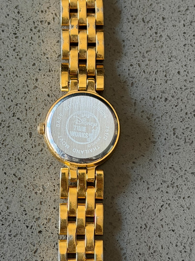 May include: A gold-coloured Disney Time Works watch with a round face and a linked strap. The watch face has a silver backing with the Disney Time Works logo and the text "Thailand Movt". The strap is made of rectangular links.