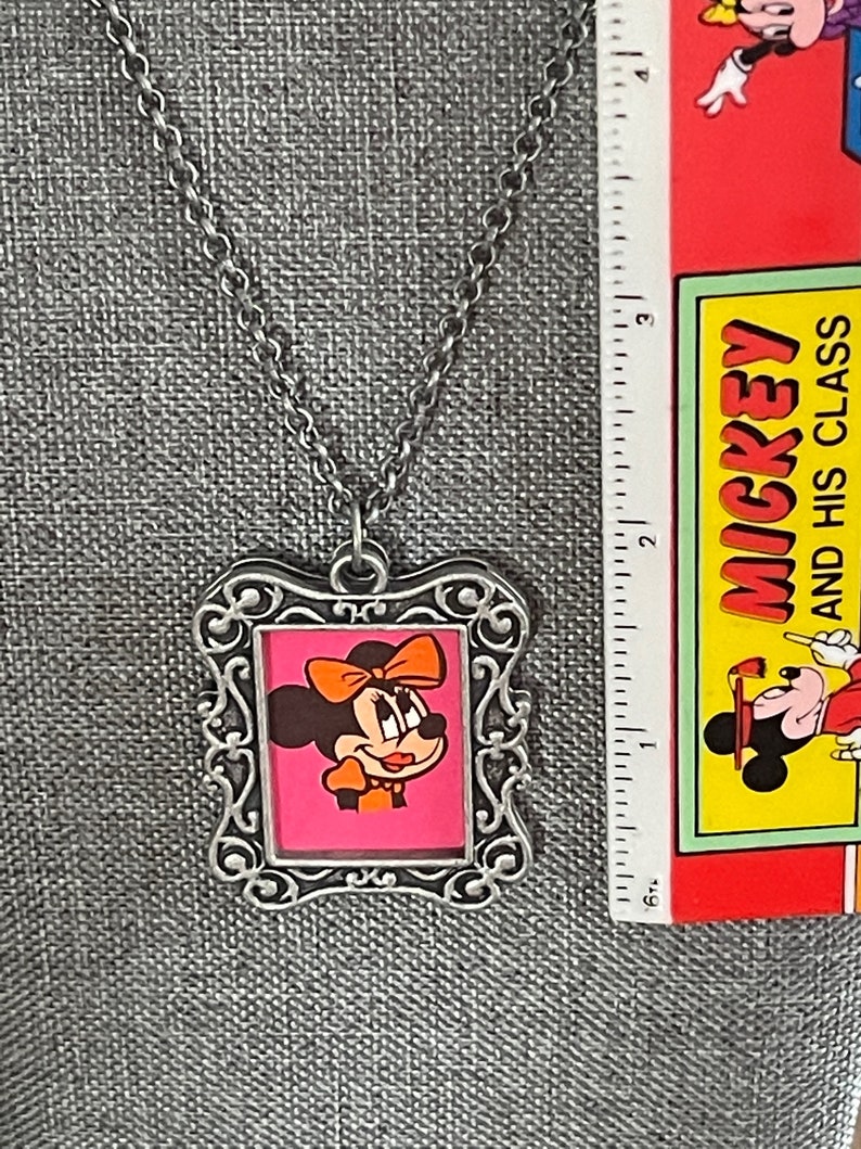 May include: Silver chain necklace with a silver picture frame pendant featuring a cartoon image of Minnie Mouse with a red bow and a pink background.