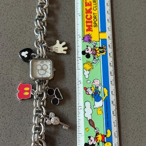 May include: Silver-toned charm bracelet watch with a square face and Mickey Mouse-themed charms. Charms include a Mickey Mouse glove, heart, and key. A ruler with Mickey Mouse characters is next to the watch.