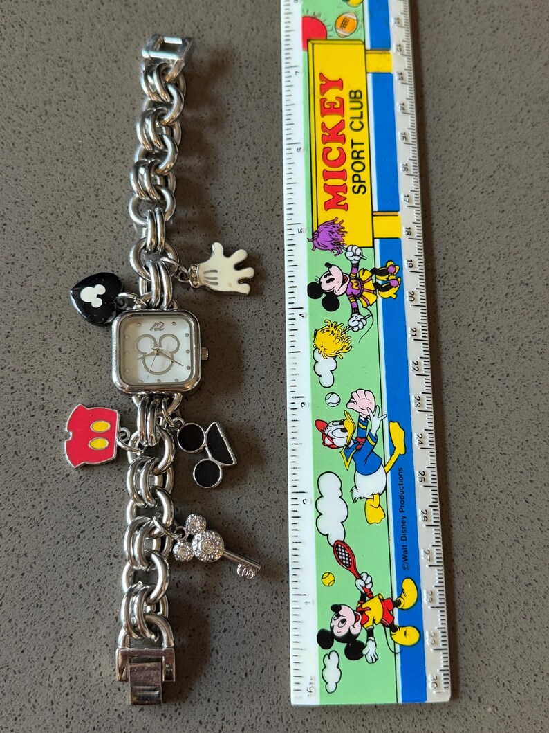 May include: Silver-toned charm bracelet watch with a square face and Mickey Mouse-themed charms. Charms include a Mickey Mouse glove, heart, and key. A ruler with Mickey Mouse characters is next to the watch.
