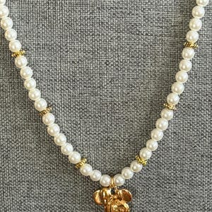 May include: A white faux pearl necklace with gold-colored accents. The necklace features a gold-colored pendant in the shape of a cartoon mouse. The necklace is displayed against a gray textured background.