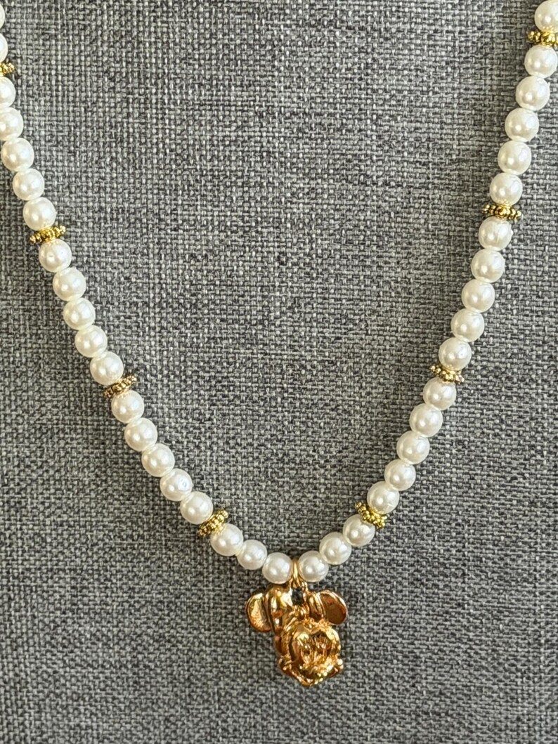 May include: A white faux pearl necklace with gold-colored accents. The necklace features a gold-colored pendant in the shape of a cartoon mouse. The necklace is displayed against a gray textured background.