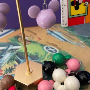 Puede incluir: Un conjunto de pendientes de Mickey Mouse de colores vivos. Los pendientes est&aacute;n hechos de pl&aacute;stico y tienen un acabado brillante. Los pendientes se muestran en un soporte dorado. Los pendientes son de varios colores, incluyendo rosa, verde, blanco, rojo y negro.