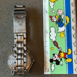 Op de afbeelding: Een roestvrijstalen horloge met gouden accenten en een Mickey Mouse-embleem op de sluiting. Het horloge ligt naast een liniaal met Disney-personage-illustraties. De horlogeband is ongeveer 18 cm lang.