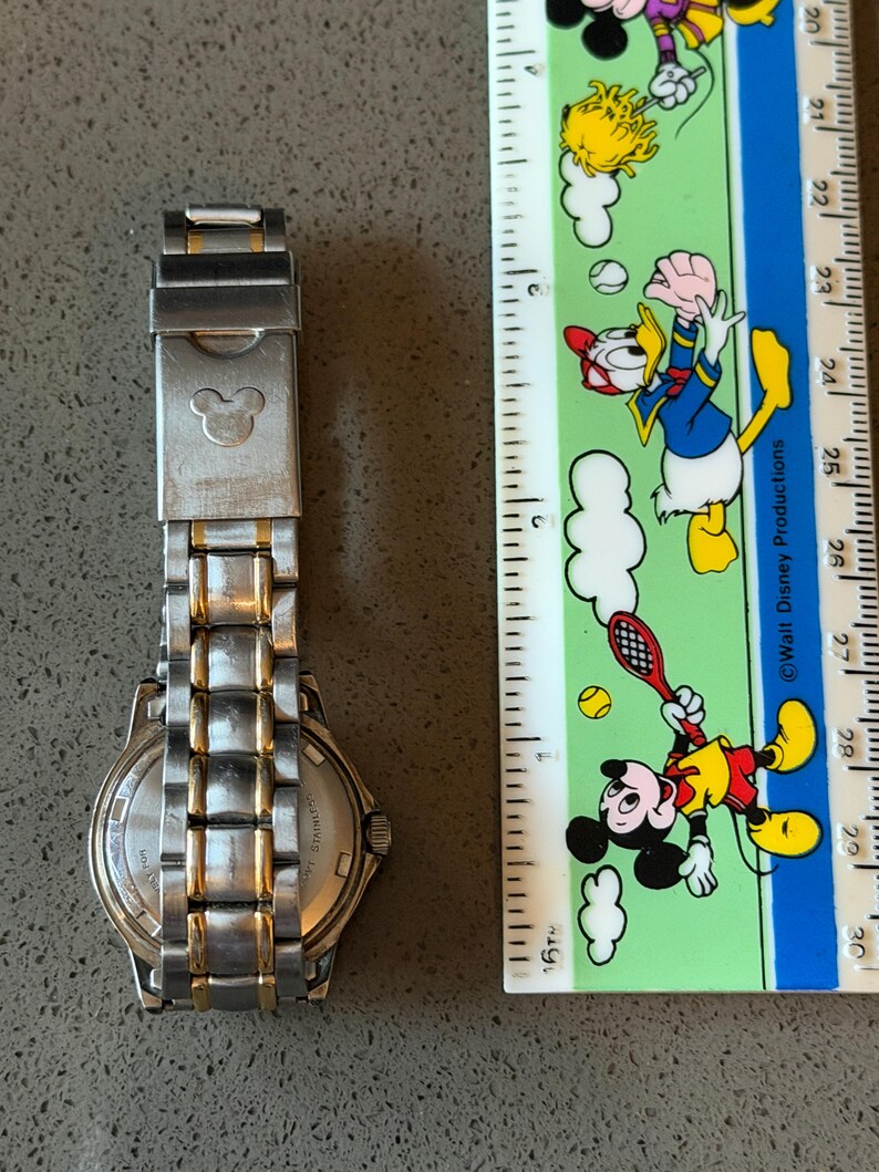 Op de afbeelding: Een roestvrijstalen horloge met gouden accenten en een Mickey Mouse-embleem op de sluiting. Het horloge ligt naast een liniaal met Disney-personage-illustraties. De horlogeband is ongeveer 18 cm lang.