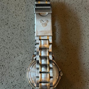 Op de afbeelding: Zilver- en goudkleurig roestvrijstalen horloge met een Mickey Mouse-silhouet op de sluiting. Het horloge heeft een ronde wijzerplaat en een schakelband. Het horloge is een klassieke stijl.