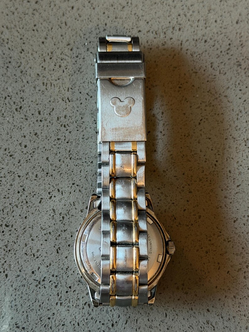 Op de afbeelding: Zilver- en goudkleurig roestvrijstalen horloge met een Mickey Mouse-silhouet op de sluiting. Het horloge heeft een ronde wijzerplaat en een schakelband. Het horloge is een klassieke stijl.