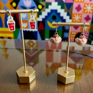 May include: Two pairs of earrings displayed on a gold stand. The first pair features a red and white popcorn bucket charm. The second pair features a brown and white seashell charm.