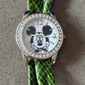 May include: A silver wristwatch with a white face featuring a Mickey Mouse design. The watch has a bezel encrusted with small, clear stones and a green, textured band. The watch face displays the numbers 12, 3, and 9.